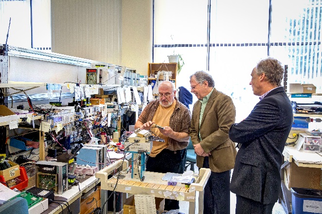 Three men standing at a work bench looking at a circuit board.